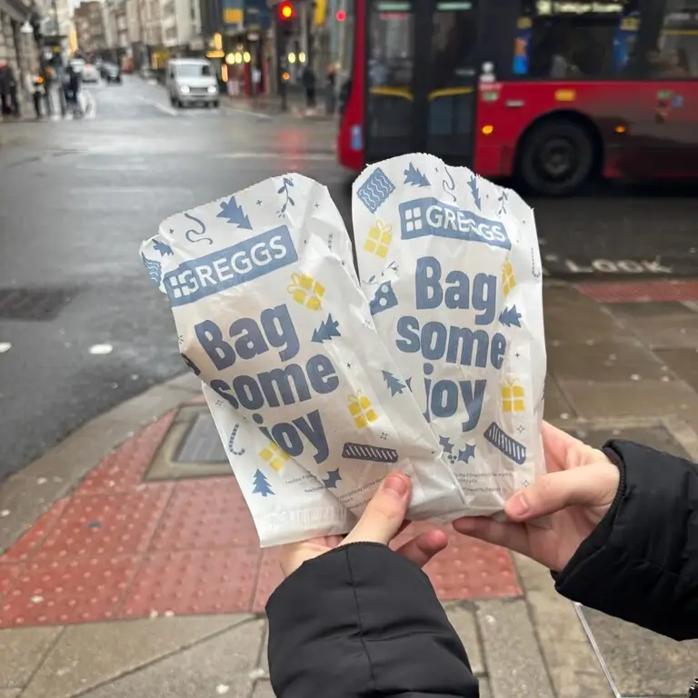 Hands holding a Greggs bag with a bustling London street in the background, capturing a moment of city life and local treats